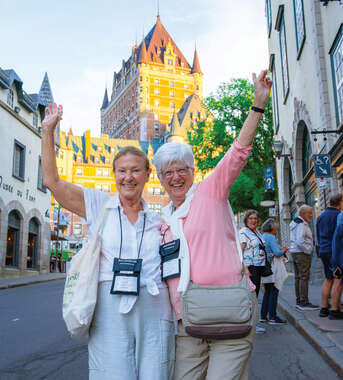 Two smiling women stand on a street in Old Québec City with arms raised, featuring the Château Frontenac in the background.