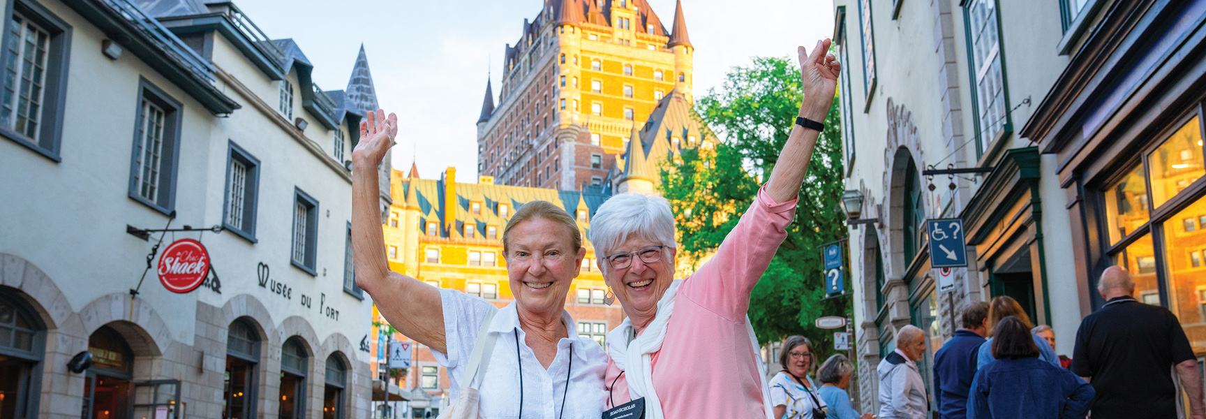 Two smiling women wave on a city street in Québec with the historic Château Frontenac hotel behind them.