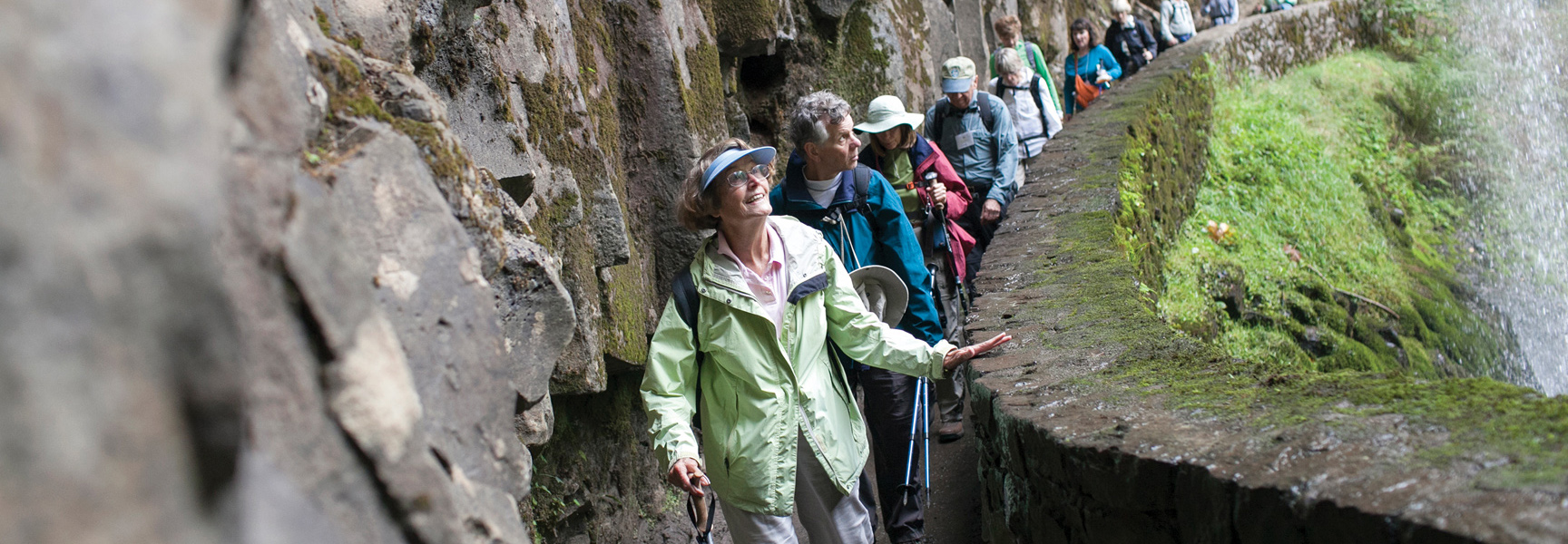 A group of hikers walk single-file on a narrow stone path carved into a rock wall next to a waterfall in Oregon.