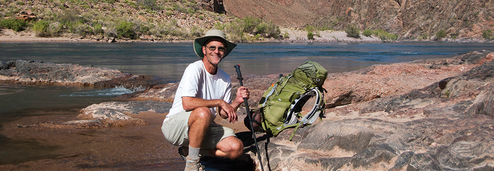 A smiling hiker crouches on the rocky shore of a river in the Grand Canyon, Arizona, with his backpack and trekking poles beside him.