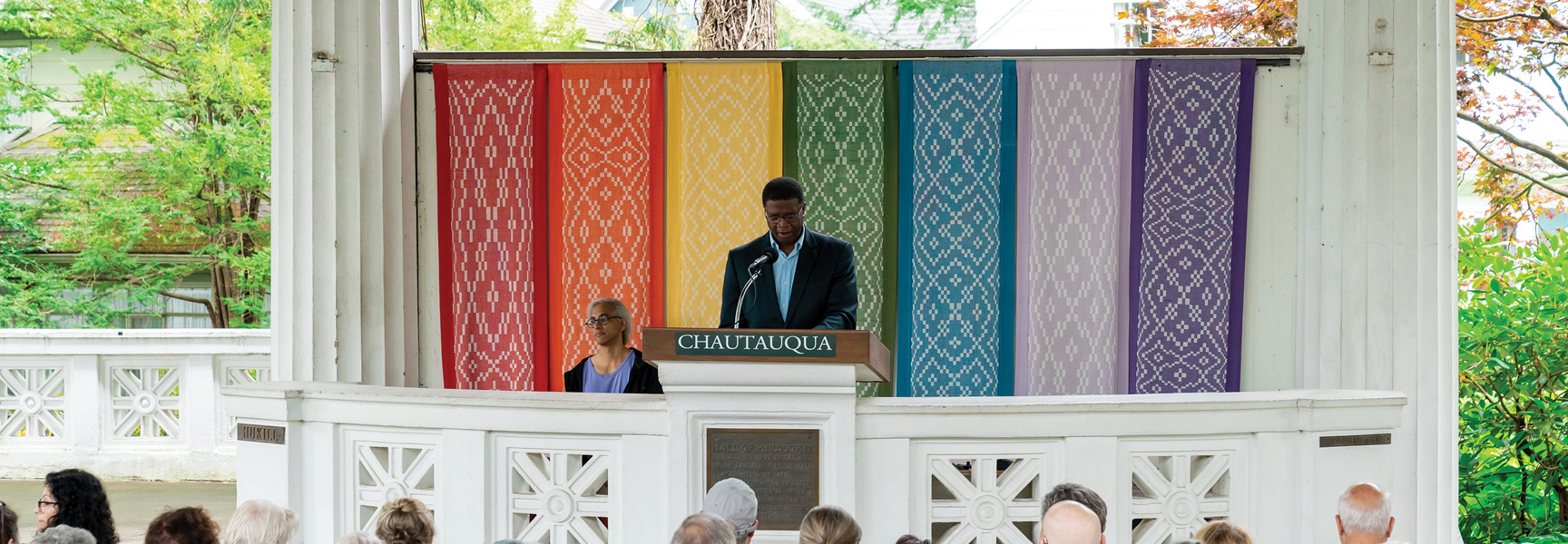 A man speaks at a Chautauqua podium in New York, with colorful banners behind him as an audience listens from below.