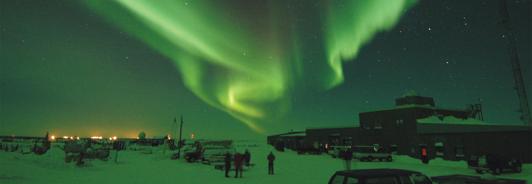 The vibrant green aurora borealis streaks across the starry night sky above a snowy landscape with buildings and people in Manitoba.