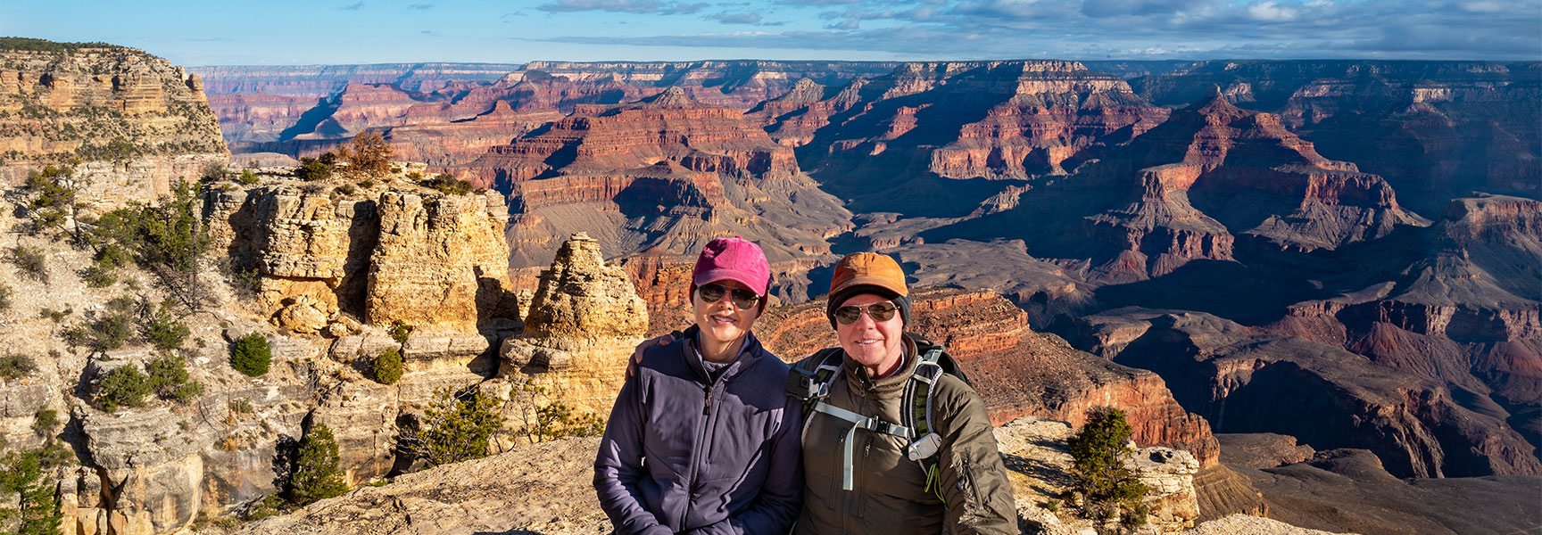 Two hikers smile at the camera from Ooh Aah Point, with the expansive, sunlit Grand Canyon in Arizona stretching out behind them.