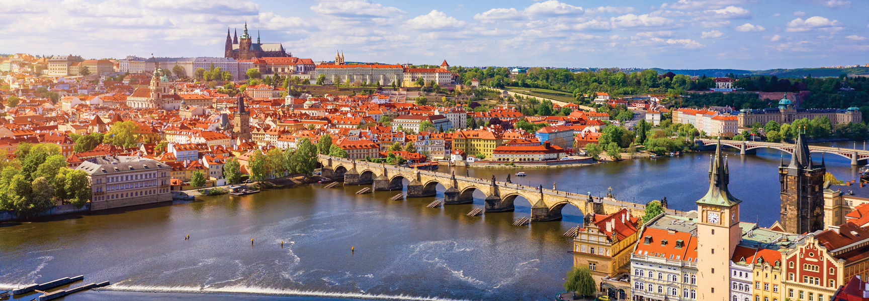 A sunny, panoramic view of Prague in the Czech Republic, showing the Charles Bridge over the Vltava River with Prague Castle in the distance.
