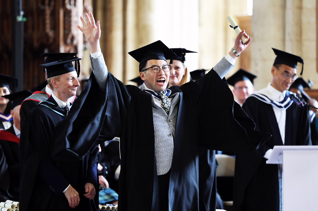 A smiling male graduate in a cap and gown raising both arms with a rolled-up scroll in one hand