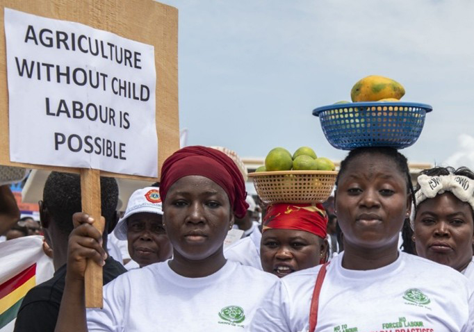A group of people are gathered for a protest or awareness march. In the foreground, a woman wearing a white t-shirt and a dark red headscarf holds a wooden sign that reads, 'AGRICULTURE WITHOUT CHILD LABOUR IS POSSIBLE.' To her right, another woman in a white t-shirt balances a blue basket filled with fruit on her head. Directly behind her, a third woman balances a smaller tan basket of green fruit.
