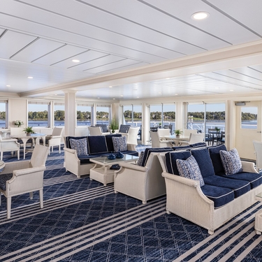 Elegant interior of an American Cruise Lines constellation-class sky lounge featuring blue and white seating with panoramic window views of the water and shoreline.