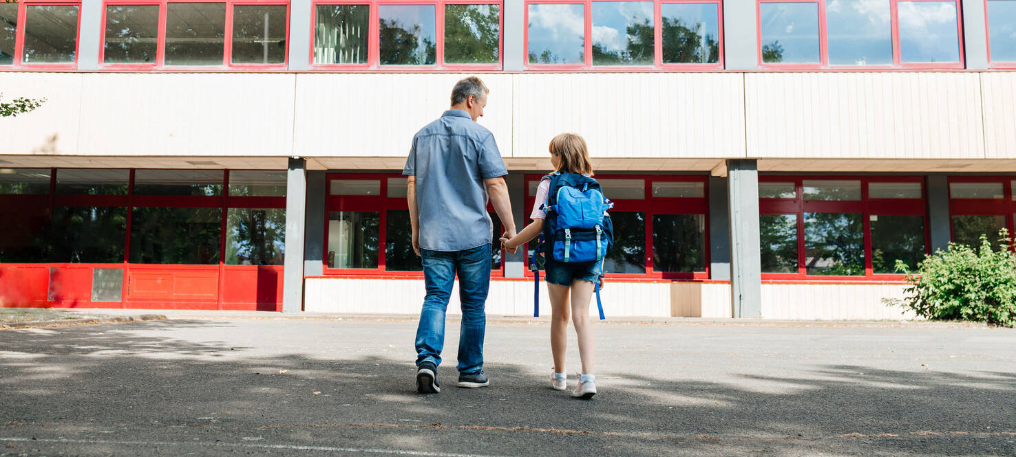 Photo shot from behind a dad holding hands with his daughter who's wearing a blue backpack and heading back to school