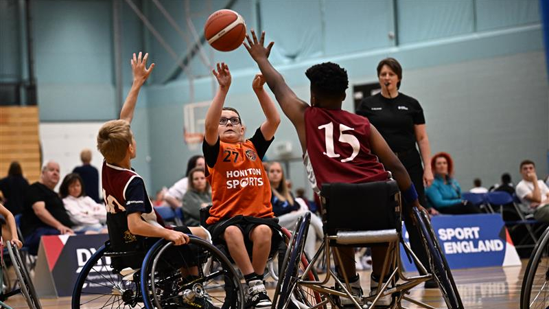 Young players in wheelchairs compete in a basketball game, with one player in an orange jersey taking a shot while two defenders raise their arms to block.