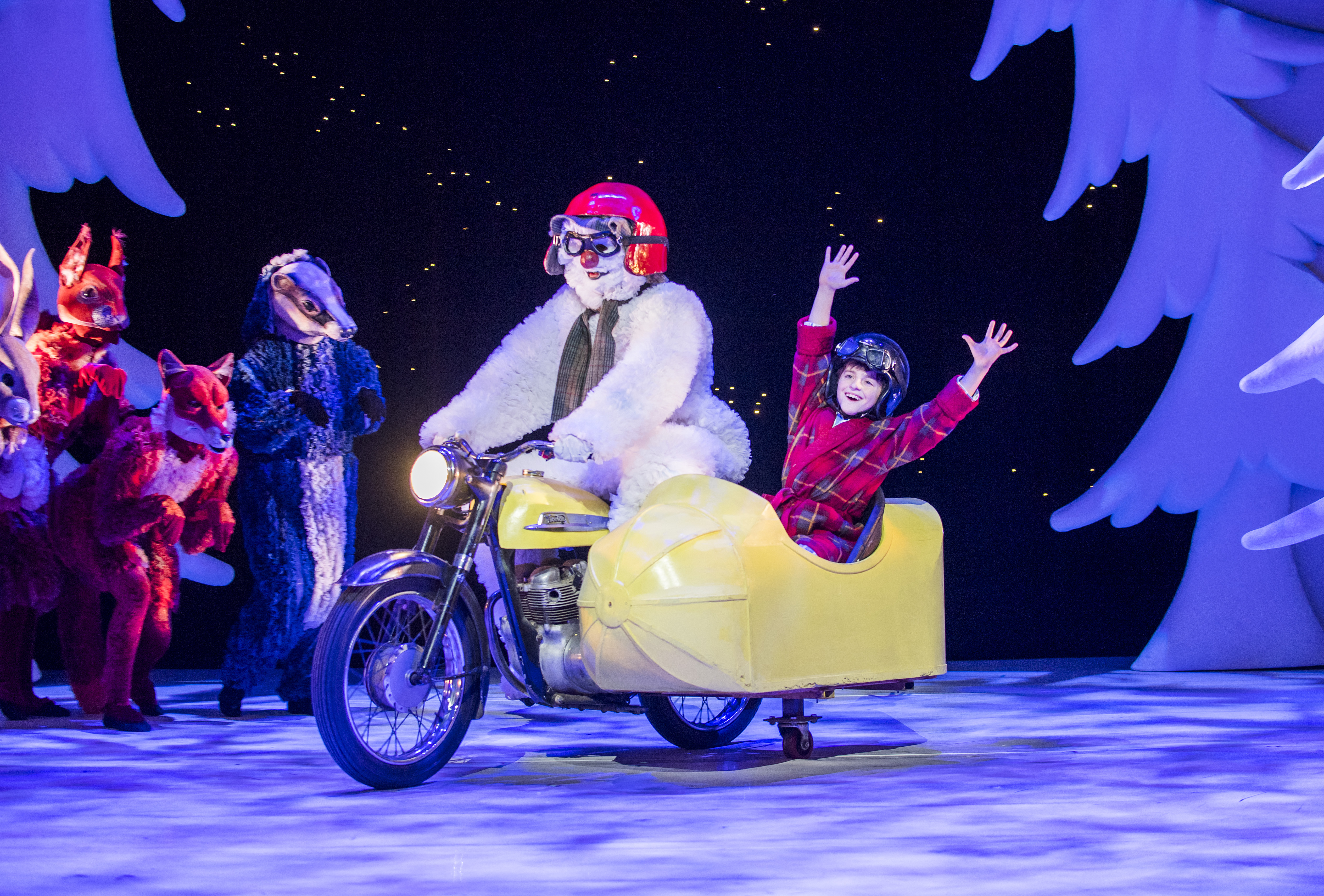 A performer dressed as a snowman rides a motorcycle with a sidecar carrying a joyful child in red pyjamas, surrounded by characters in animal costumes on a snowy stage.