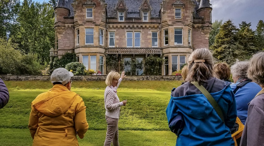 A woman points to the Aigas building in the background while speaking to a group