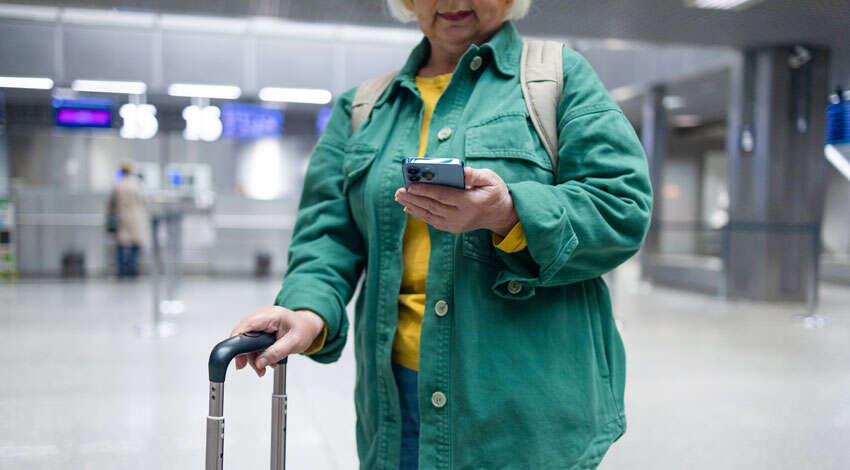 A woman in a green jacket looks at her smartphone while holding a suitcase handle in an airport terminal.