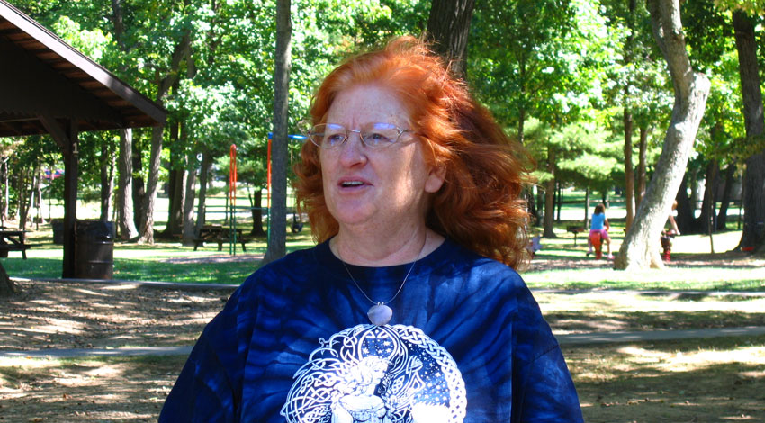 A woman with bright red hair and glasses, wearing a blue patterned t-shirt, stands in a park with trees in the background.