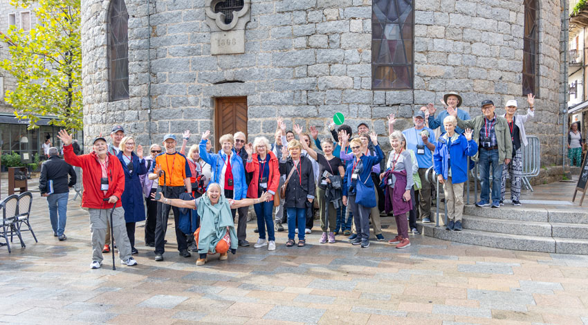 A large group of travelers smiles and waves for a group photo in a stone plaza in Corsica.