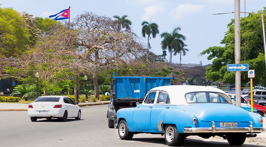 A blue classic car drives on a street with the Cuban flag in the background.