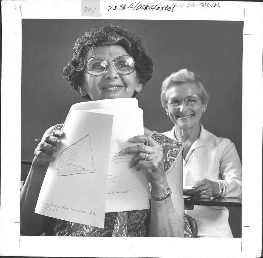 Two women smile at the camera, and the woman in the foreground holds up papers showing her work from class