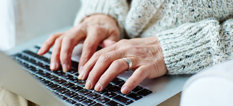 An older adult wearing a knit sweater uses a laptop, likely researching financial assistance options.