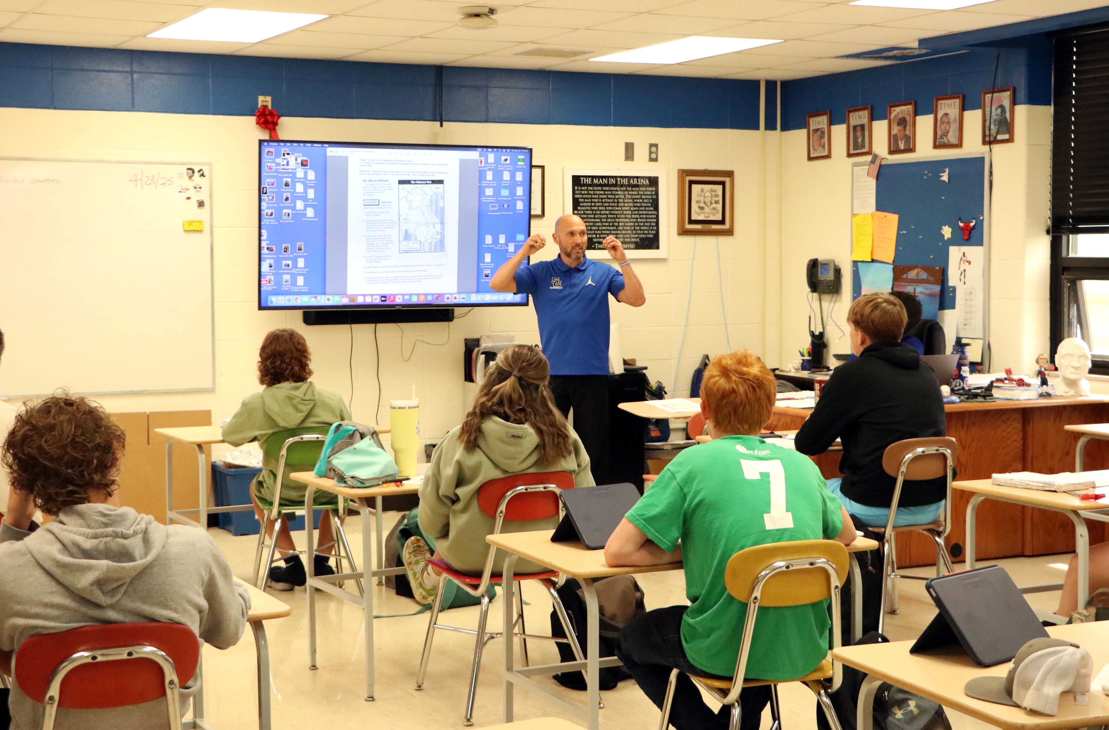 Christopher Gray teaches students in a classroom while standing next to a large screen displaying a history lesson.