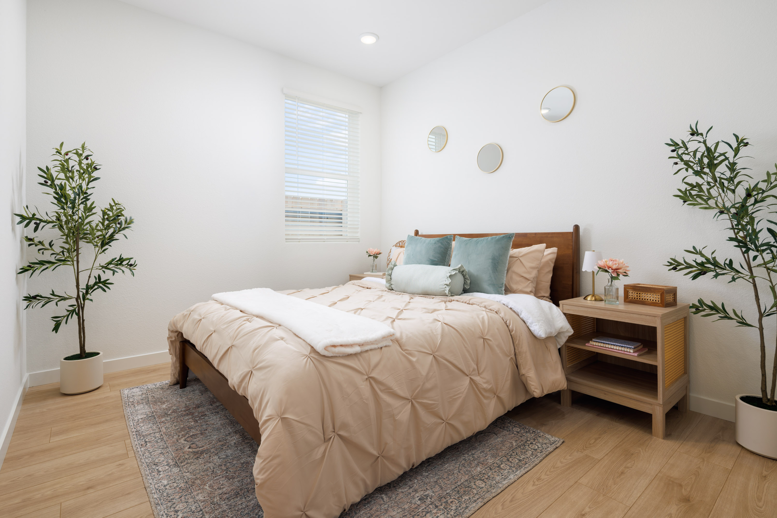 bedroom with wood floors, two potted olive plants, and tan bedding with two aqua velvet pillows