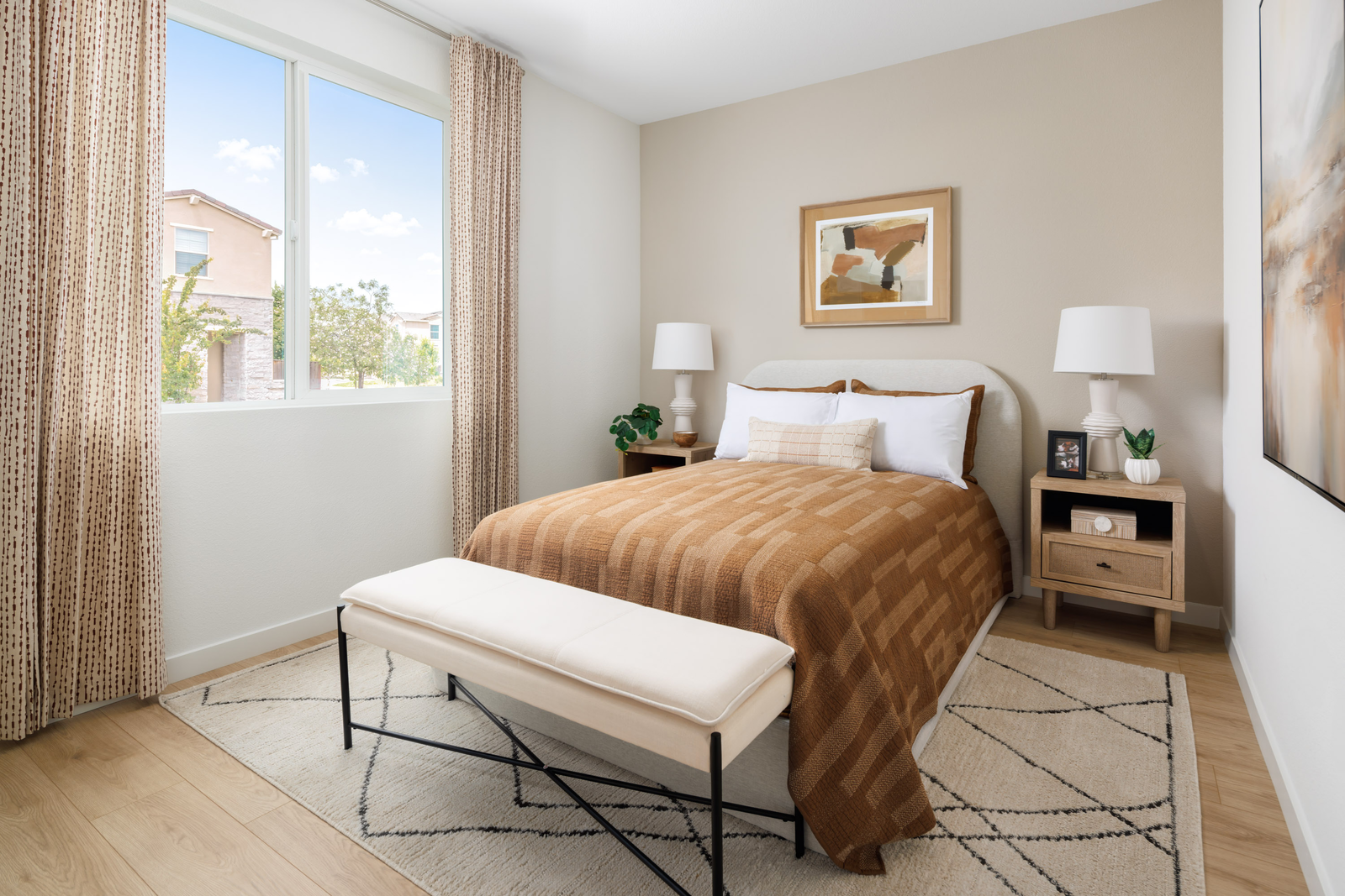 bedroom with large window, tan upholstered bed with brown quit, and light wood floors