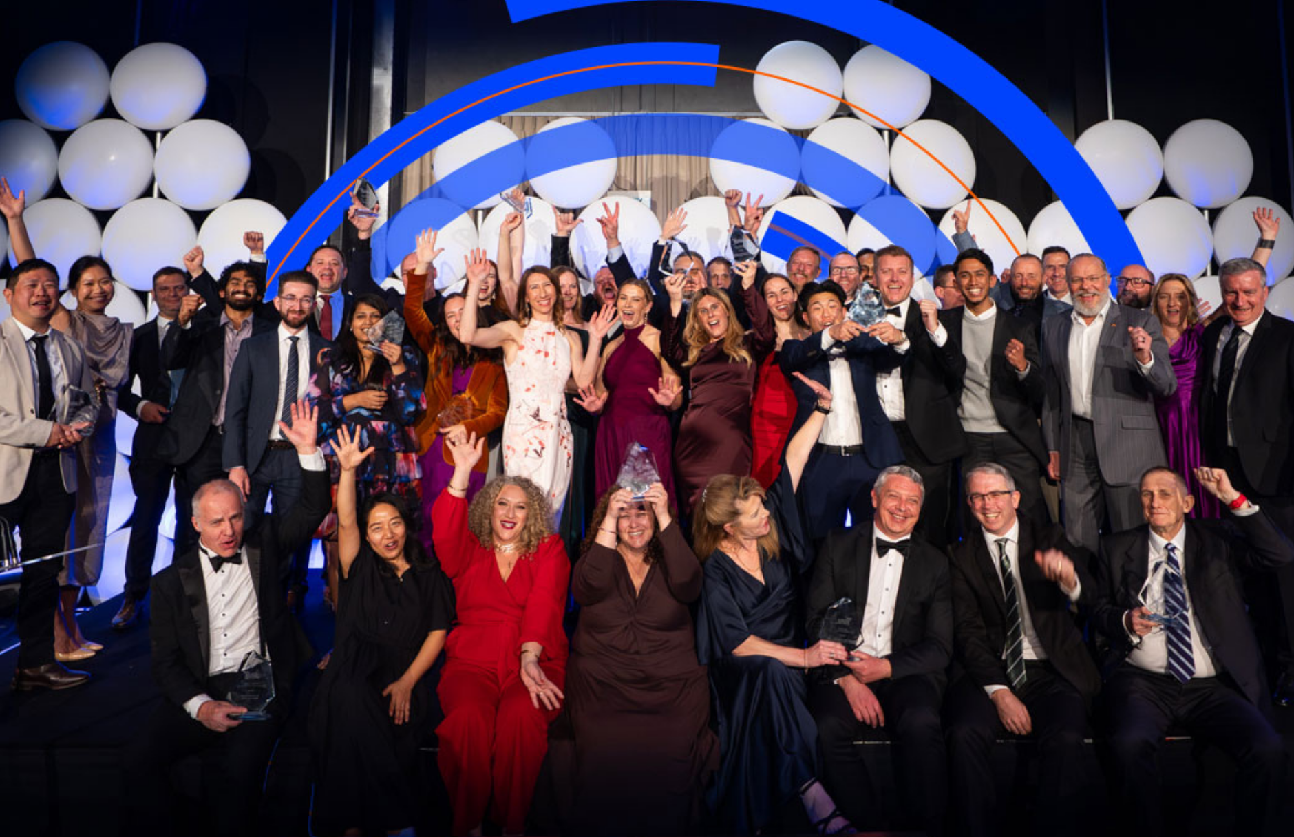 A large group of people celebrate on a stage at the CIPS ANZ Excellence in Procurement Awards. Many people in the group are cheering with their arms raised, and some are holding glass trophies. The people are dressed in formal attire, including suits, tuxedos, and evening dresses. The background features a wall of large white balloons and a blue circular graphic.
