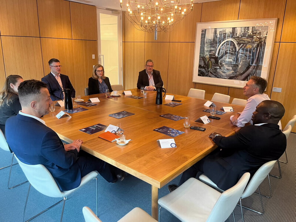 A group of business professionals, four men and two women, are seated around a large square wooden table during a CPO (Chief Procurement Officer) roundtable meeting. The table is set with professional documents and coffee. A large painting of a cityscape is on the wall.