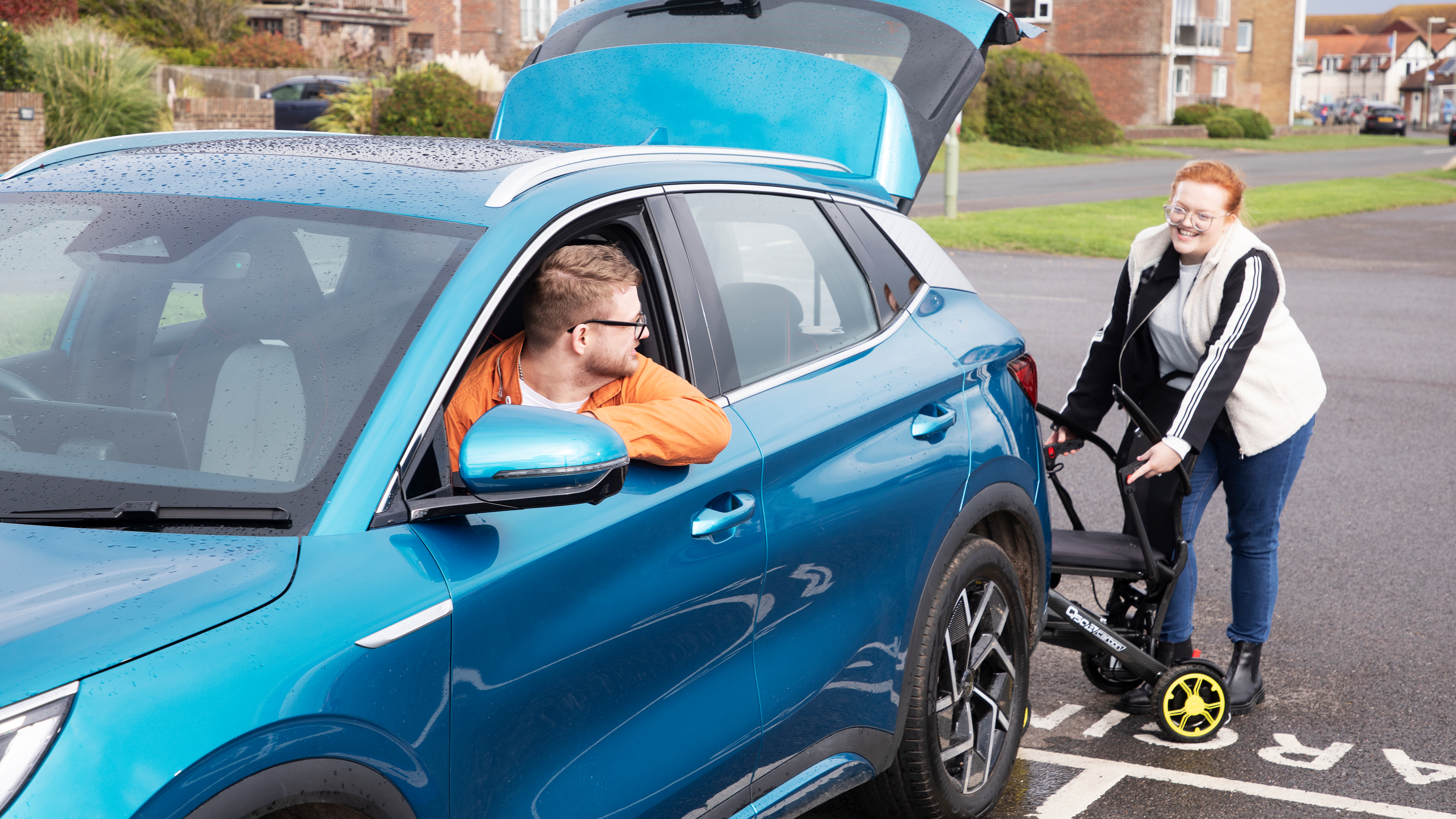 A woman loads a folded mobility scooter into the boot of a blue SUV while a man sits in the driver’s seat looking back at her. The car is parked on a residential street with the tailgate open.