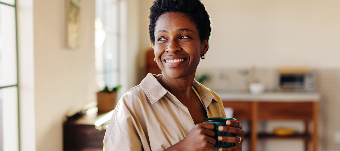 A Black woman smiles out of her window while drinking out of a coffee mug