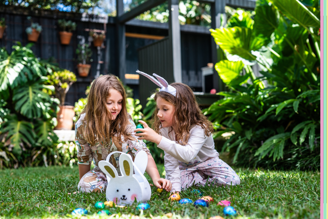 Two children in Easter themed pyjamas, one wearing bunny ears, are sitting on grass examining colorful Easter eggs near a bunny-shaped basket. Lush plants and hanging pots are in the background.