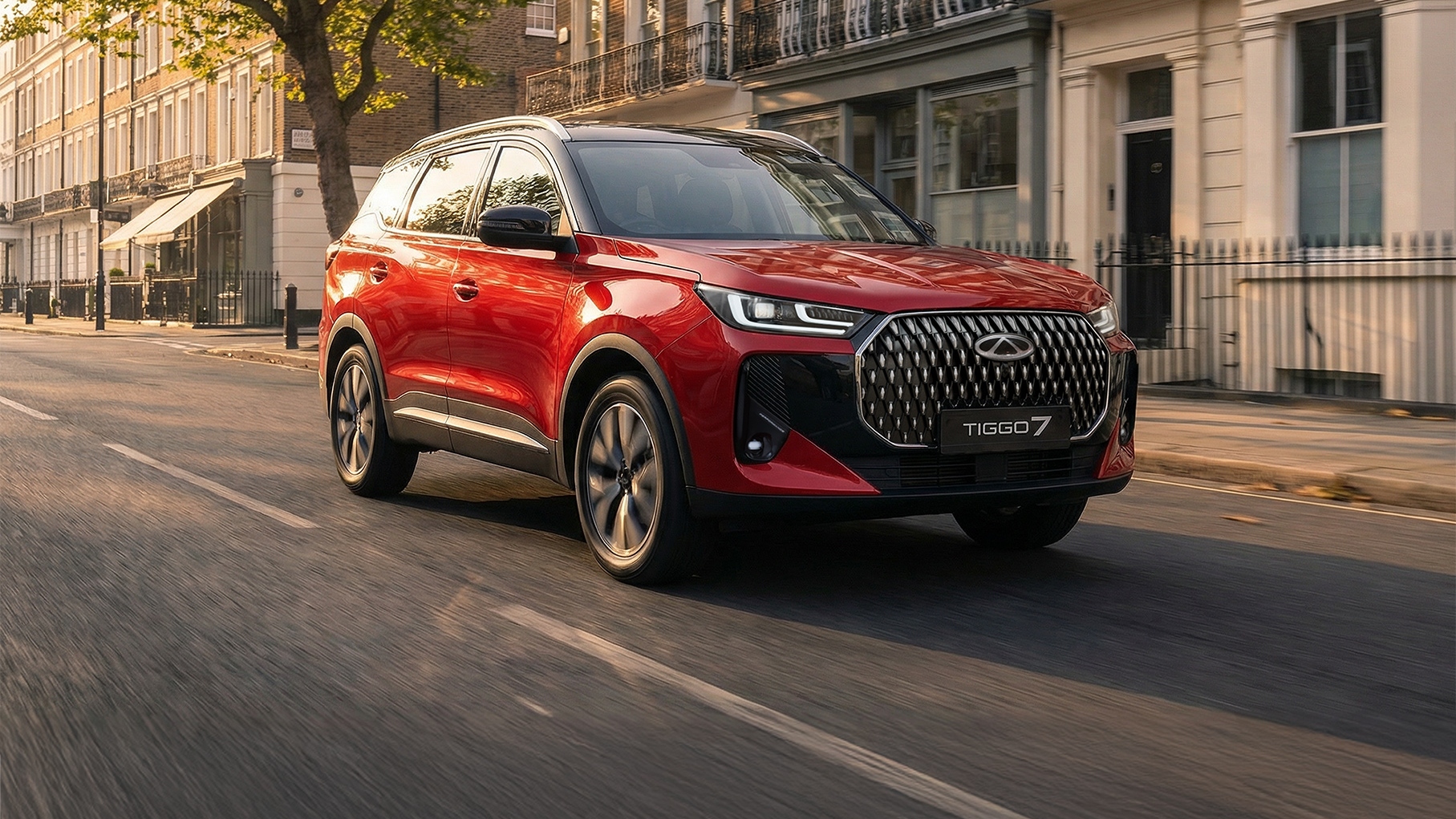 Red Chery Tiggo 7 SUV driving along a sunlit city street lined with townhouses and trees, shown from the front three quarter angle.