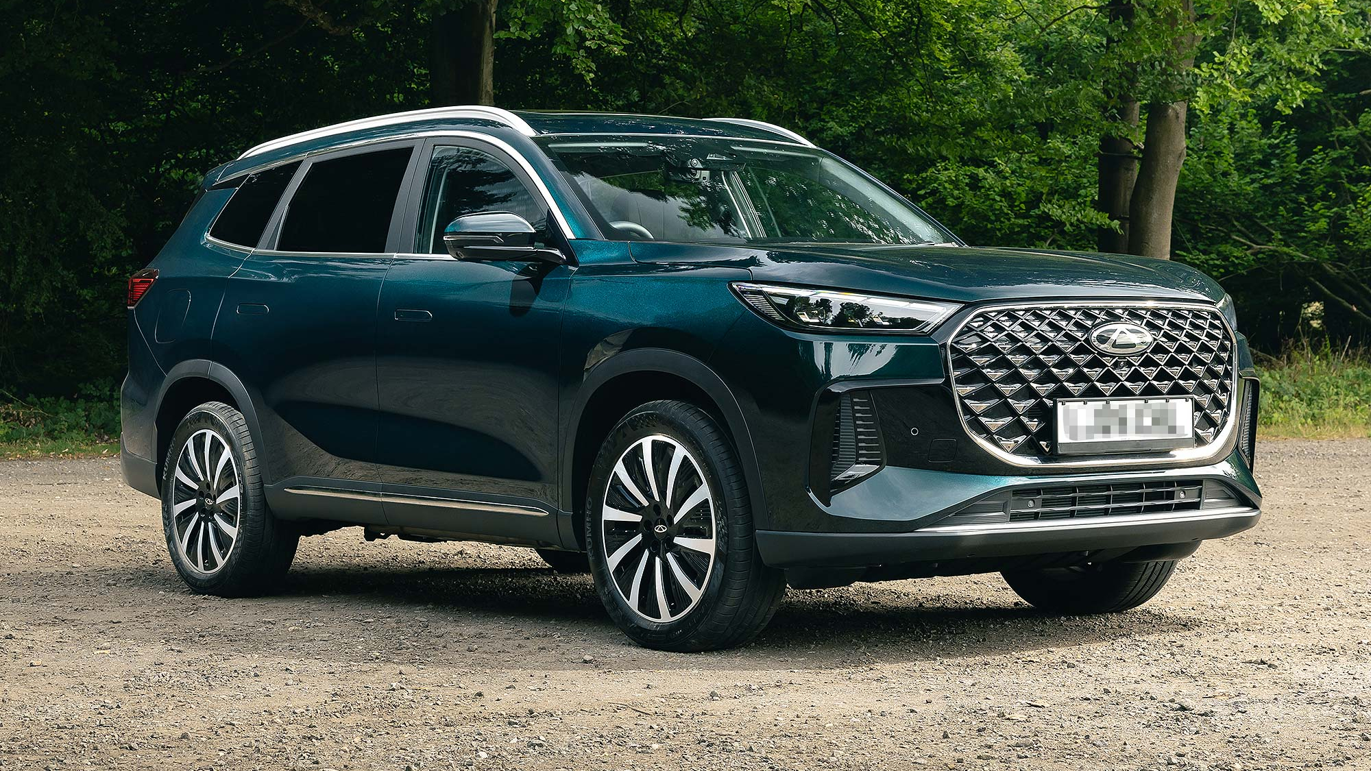 Dark green Chery Tiggo 8 parked on a gravel area beside woodland, front three quarter view showing large grille and alloy wheels.
