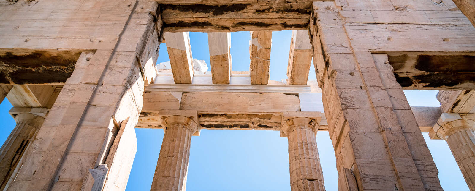 A low-angle view of the ancient stone columns and architectural beams of the Acropolis in Athens against a clear blue sky.