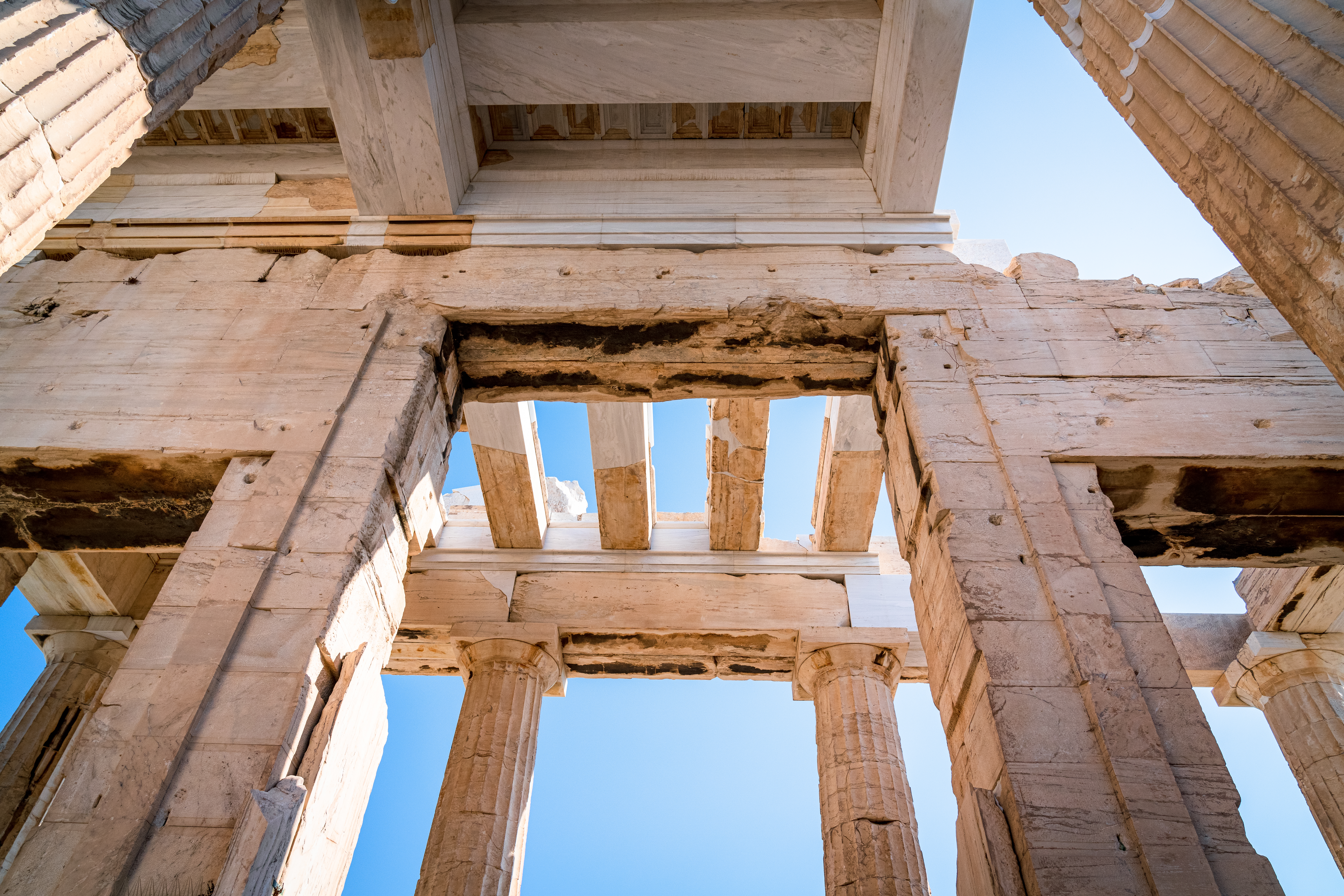 A low-angle view of the ancient stone columns and architectural beams of the Acropolis in Athens against a clear blue sky.