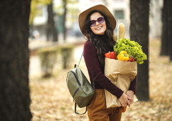 Lächelnde Frau mit Hut und Sonnenbrille trägt eine Tüte Obst und Gemüse