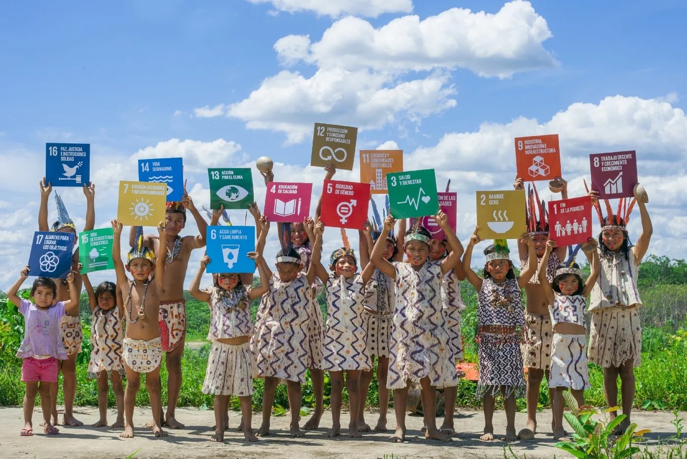 A group of children in traditional indigenous clothing holding up colorful signs representing the UN Sustainable Development Goals against a bright blue sky.