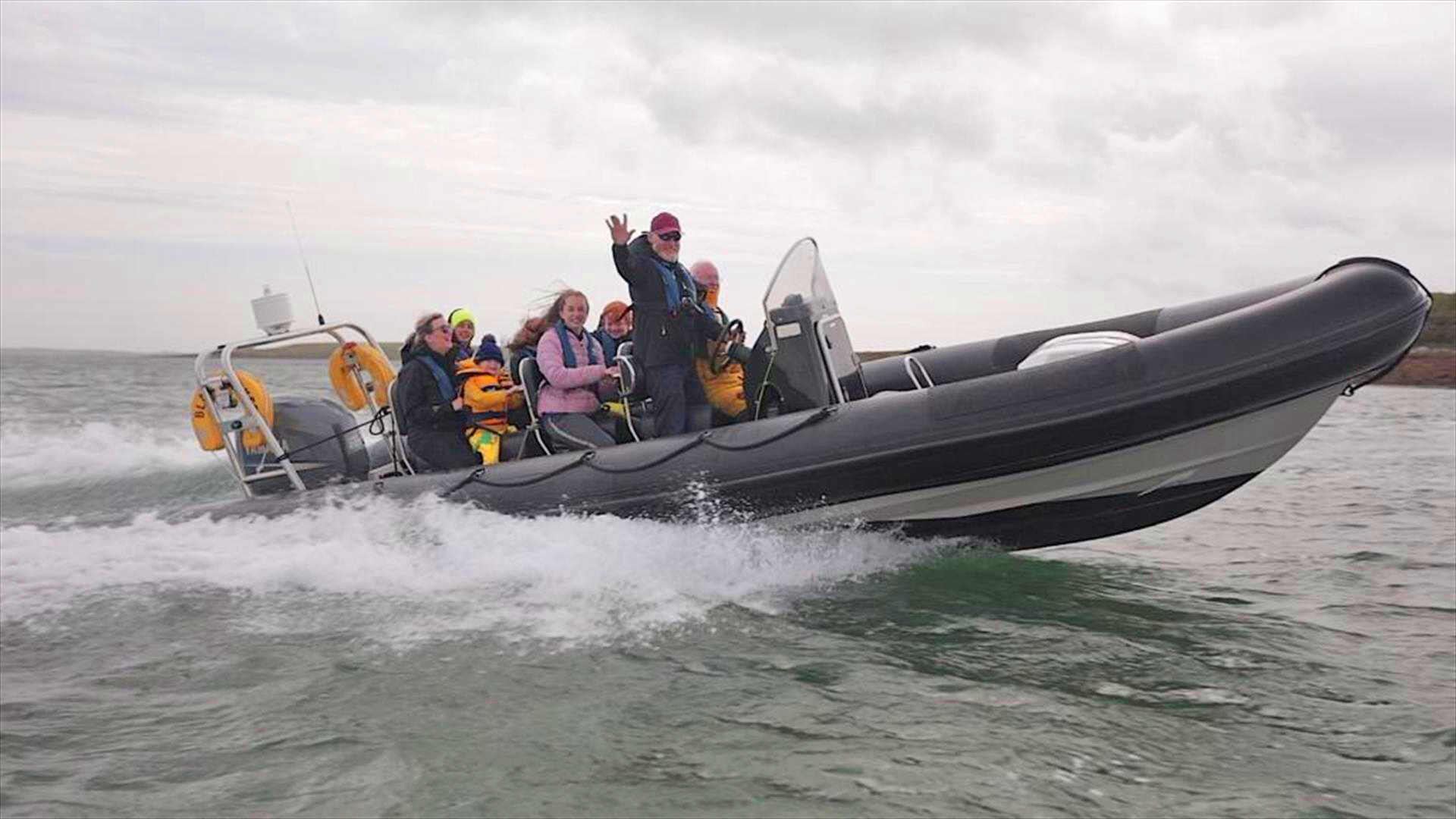 A group of people wearing life jackets ride in a fast black inflatable boat on the sea, smiling and waving as water sprays around them.