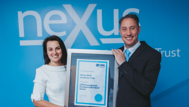Two people, a woman in a white dress and a man in a dark suit with a light blue tie, stand together holding a large, silver-framed certificate. The certificate is awarded to Nexus Multi Academy Trust for achieving the CIPS Procurement Excellence Standard Award. They are standing in front of a bright blue wall featuring a large white 'nexus' logo.