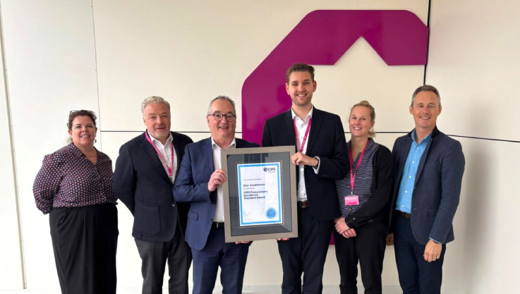 A group of six professionals stand in a line against a white wall with a large, stylized purple logo. Two men in the center hold a framed certificate awarded to Star Academies for the CIPS Procurement Excellence Standard Award. Several people in the group are wearing pink lanyards.