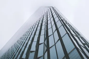 A dramatic, low-angle perspective looking up at the glass facade of a modern skyscraper that tapers into a cloudy or foggy sky.
