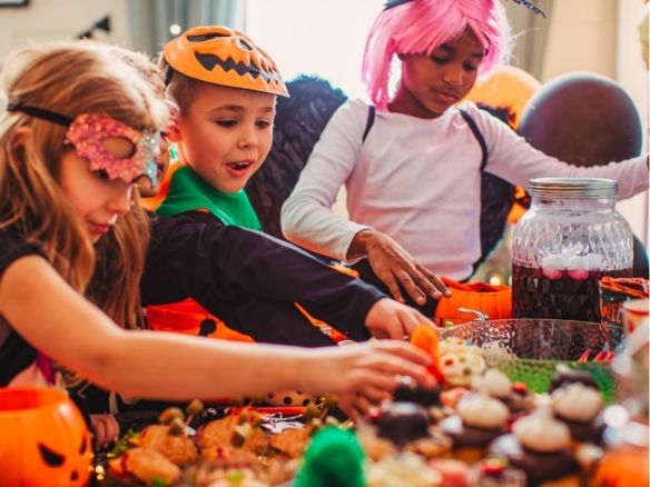 Three children in Halloween costumes gather around a table filled with colorful treats, reaching for food and candy.