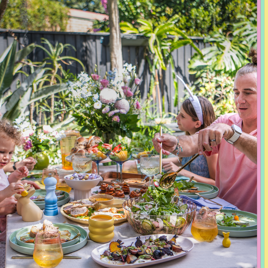 Family celebrating Easter festivities dining outdoors at a table filled with various dishes, salads, and beverages. Adults and children are engaging and serving food. Lush greenery and flowers in the background and a table adorned with Easter decorations.