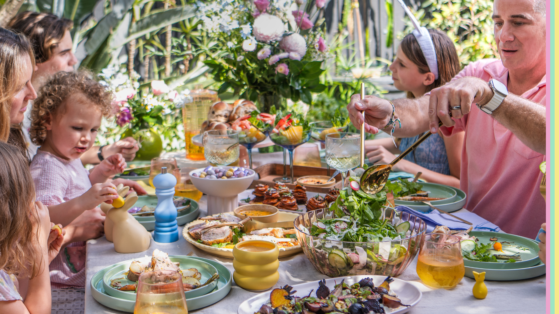Family celebrating Easter festivities dining outdoors at a table filled with various dishes, salads, and beverages. Adults and children are engaging and serving food. Lush greenery and flowers in the background and a table adorned with Easter decorations.