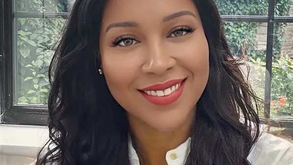 Headshot of Fayola-Maria Jack, CEO of Resolutiion, smiling warmly at the camera. She has long, dark, wavy hair, medium-toned skin, and light-colored eyes. She is wearing a light-colored top, possibly a white collared shirt, and a small stud earring. She is standing indoors in front of a window overlooking a lush green garden area.