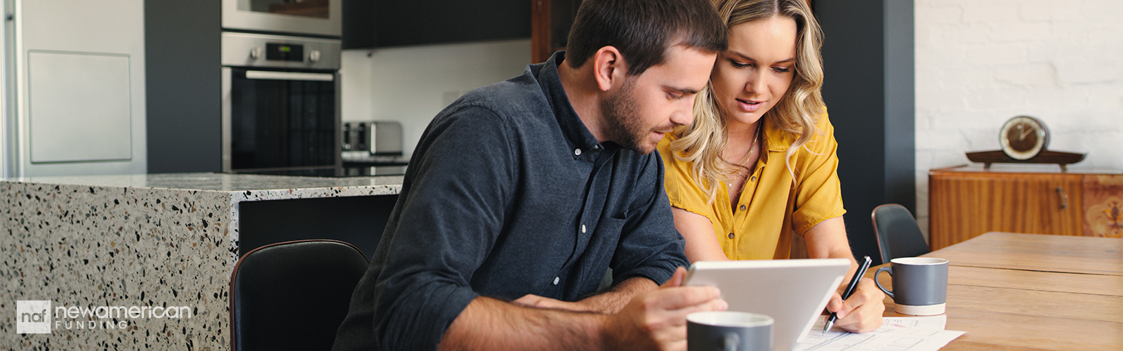 A white man and woman sit at a table looking at a tablet and writing on a piece of paper