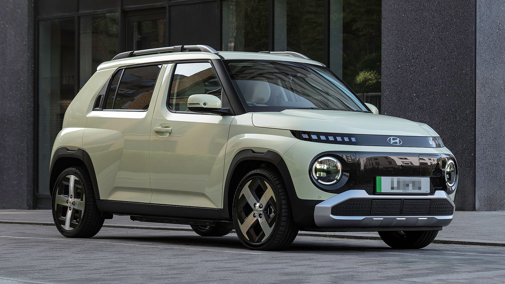 A pale green Hyundai Inster parked in an urban setting, featuring a boxy design with round headlights and black trim accents.