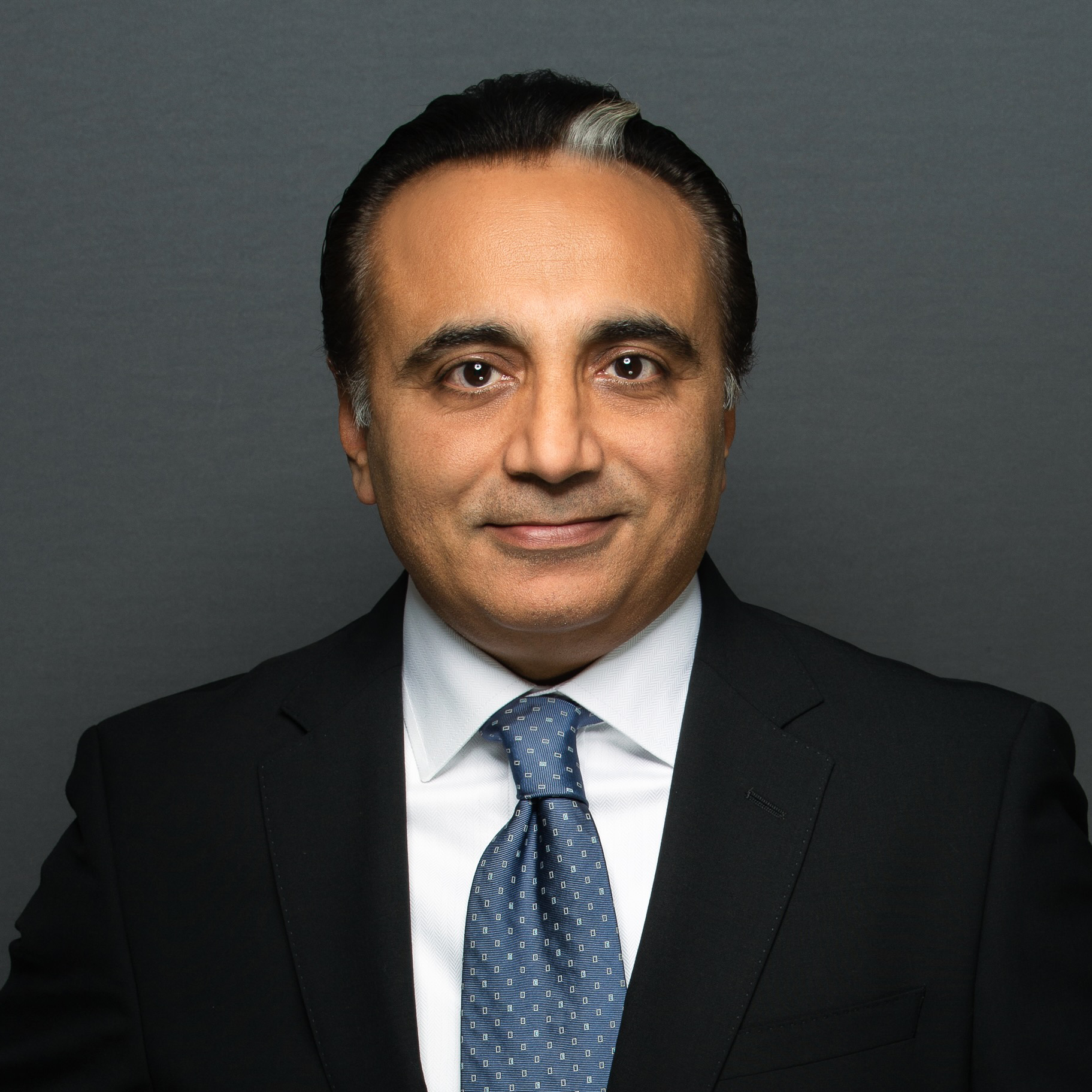 A professional studio headshot of a man with dark hair and a white streak at the temple. He is wearing a black suit, a crisp white shirt, and a blue patterned tie against a solid white background.