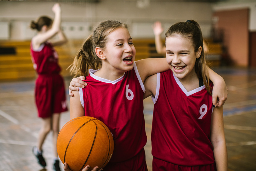Dve deklici v rdečih košarkarskih uniformah stojita objeti v telovadnici pri obšolski dejavnosti.