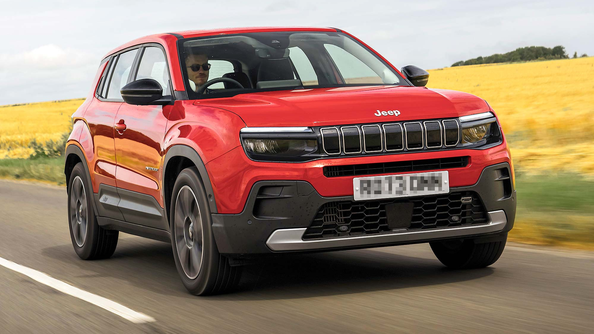 Red Jeep Avenger driving on a country road, front three quarter view, with open fields in the background.