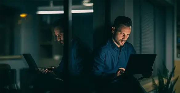 Man sat looking at a laptop screen, in a dark office room with his reflection showing in the window