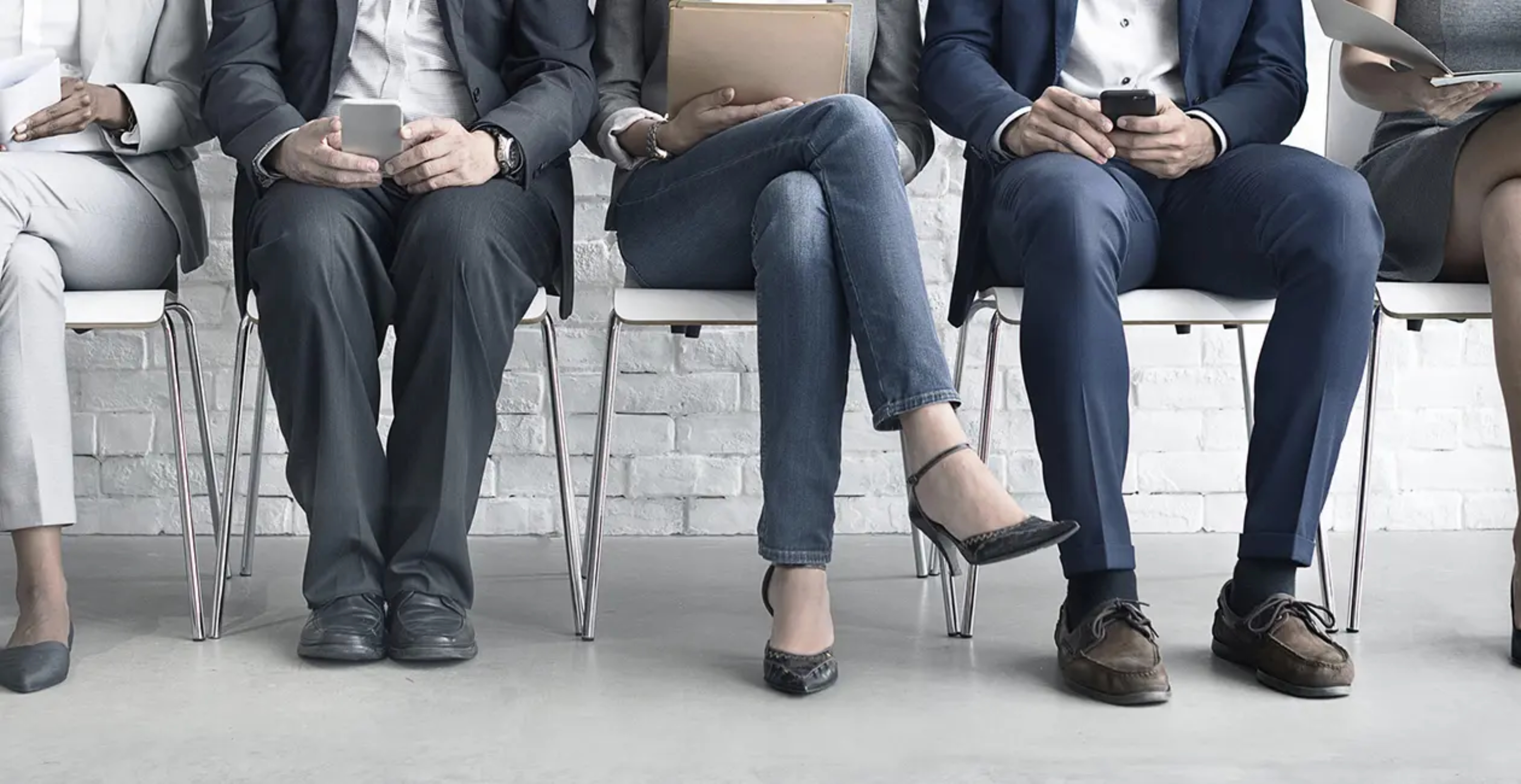 Shows a row of 5 peoples legs and torsos sat on white chairs, all wearing business attire and holding either papers or a phone
